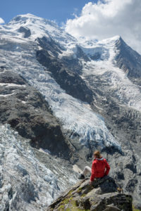 Personnage avec veste rouge assis sur un bloc de rocher devant un glacier et le Dome du Gouter à la Jonction. Massif du Mont-Blanc.